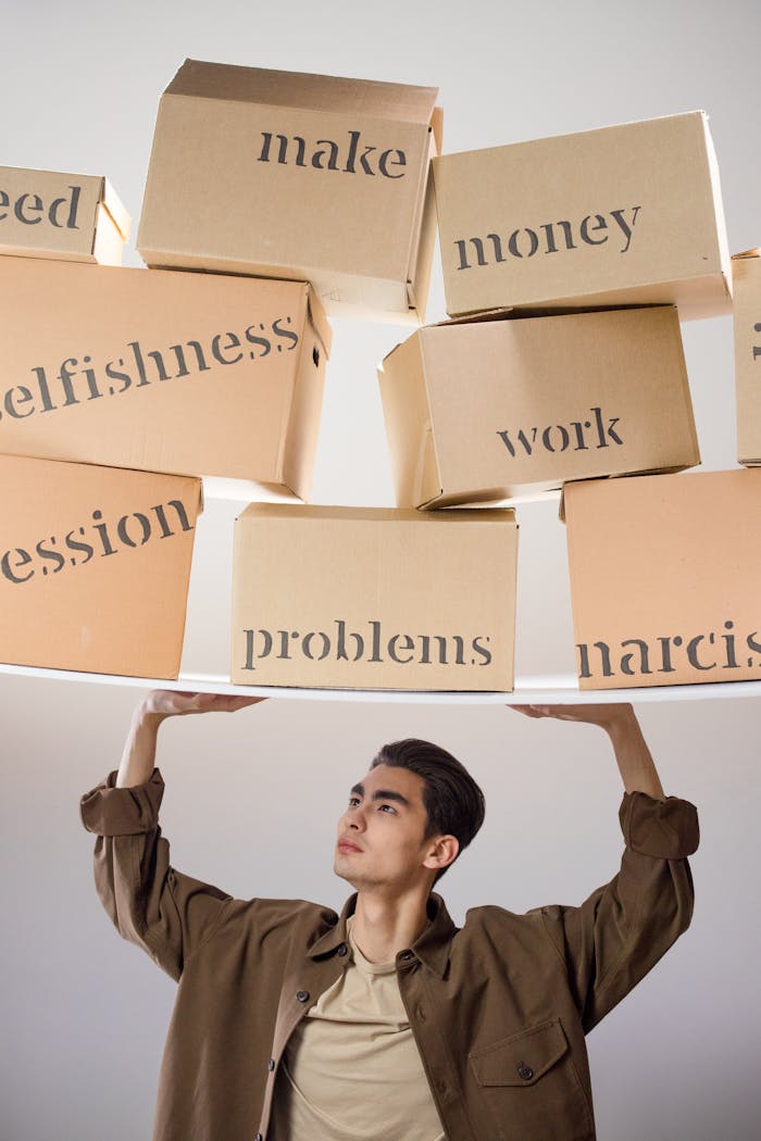 A man in a brown coat balances labeled boxes depicting various life challenges like work and money.